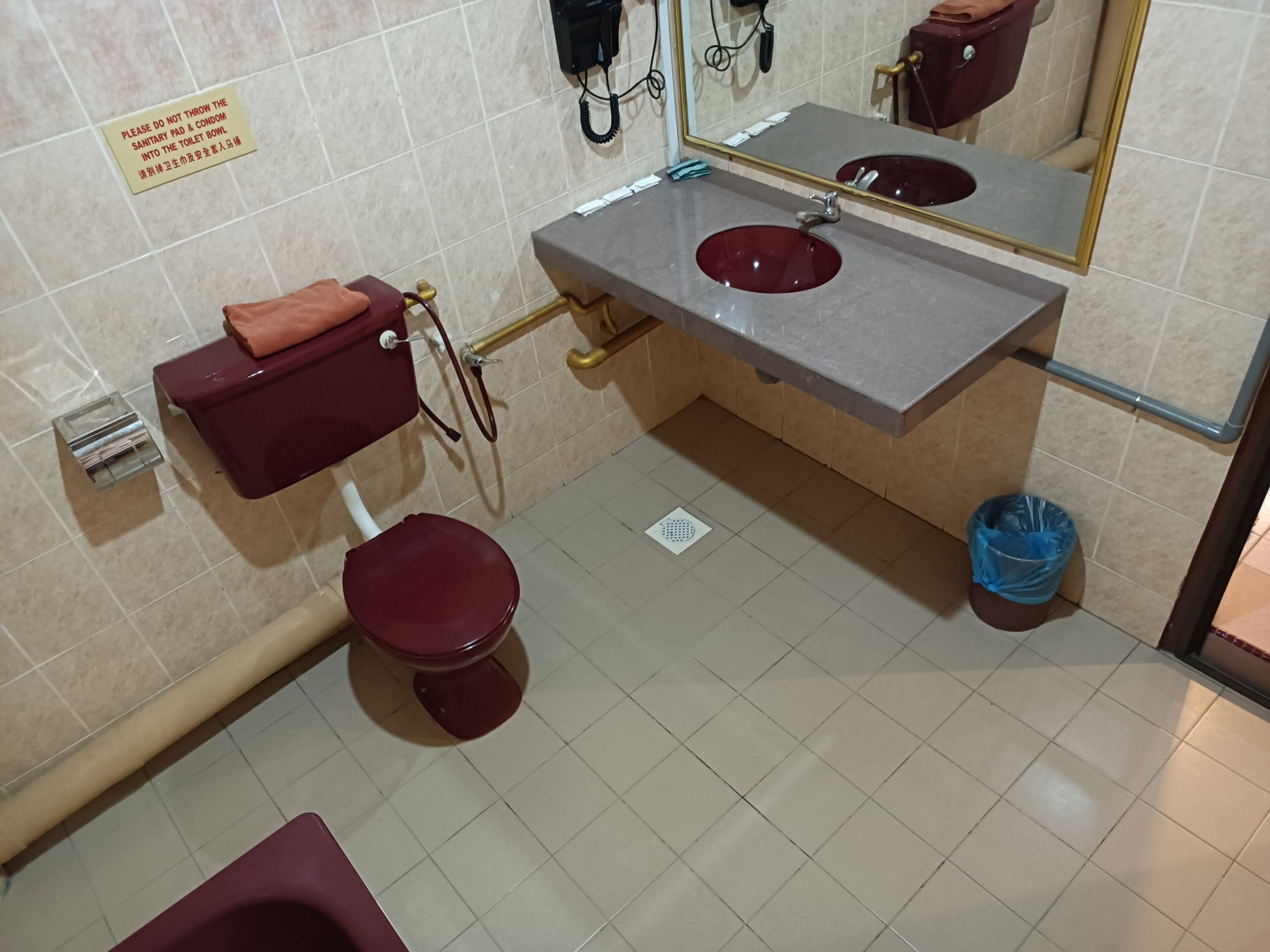A bathroom with a toilet, sink, and mirror. There is a sign on the wall and a trash bin.