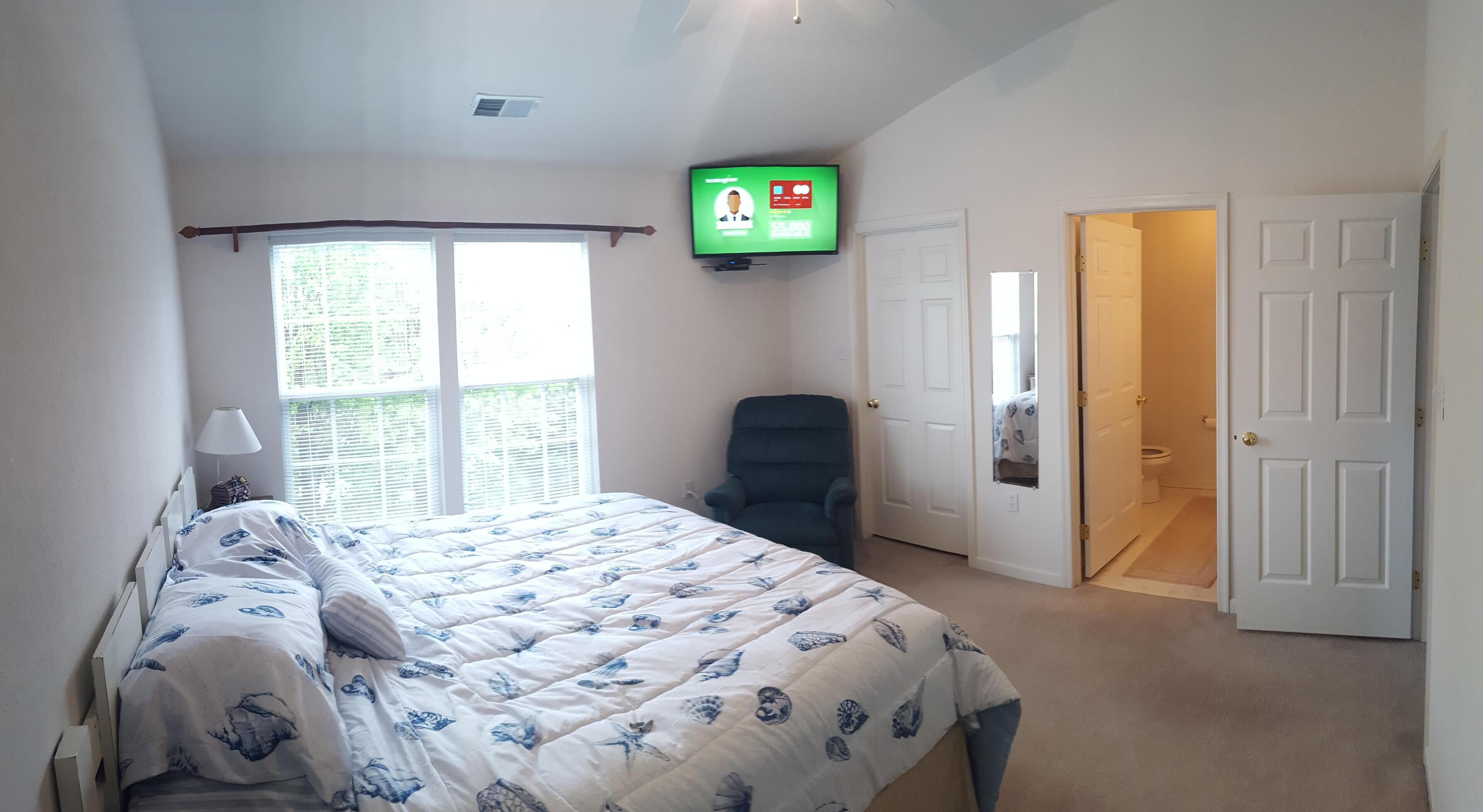 A bedroom with a bed, a chair, a television mounted on the wall, and a door leading to a bathroom.