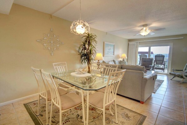 A dining area with a glass table, white chairs, a decorative wall piece, a potted plant, and a view through a glass door to the outside.