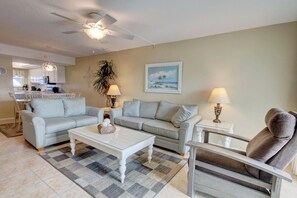 A living room with a sofa, armchair, coffee table, and lamps. There is a ceiling fan, a painting, and a plant.
