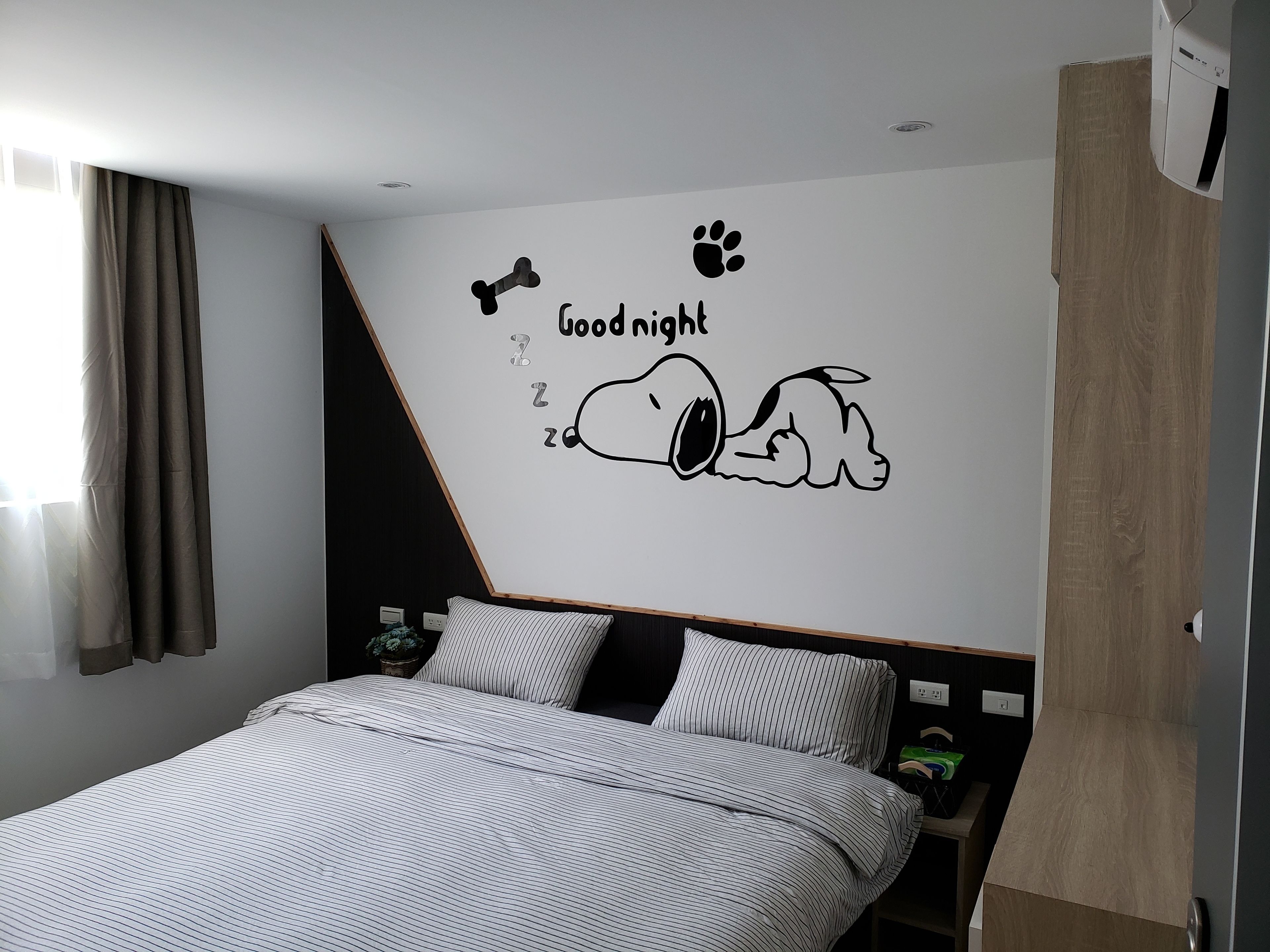 Makuuhuoneessa on sänky, seinällä on Snoopy-aiheinen seinätarra, jossa hän nukkuu, ikkunassa on verhot ja yöpöydällä on lamppu.