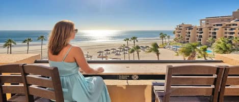 Eine Frau sitzt an einem Tisch und blickt auf einen Strand mit Palmen und einem Pool. Im Hintergrund sind Hotelgebäude zu sehen.
