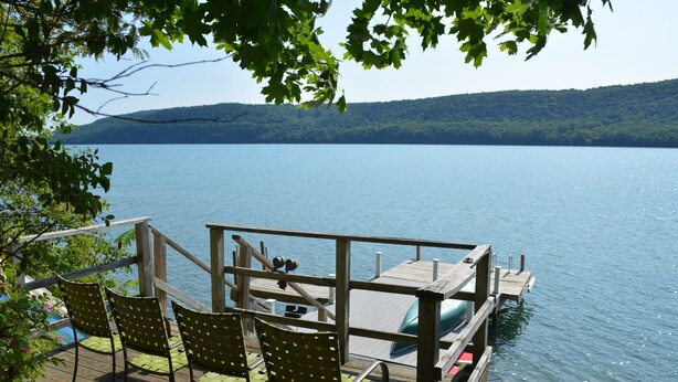 Une terrasse en bois avec des chaises donnant sur un lac, une jetée s’avançant dans l’eau et, au loin, une colline boisée.