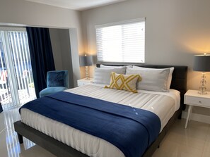 A modern hotel bedroom with a large bed, two bedside tables with lamps, a blue chair, and a view of the outdoors through a sliding door. - Luxury Rental AP3 - 3 MINUTES WALK TO THE BEACH (Pompano Beach)