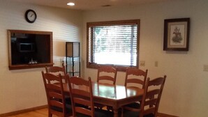 Una sala da pranzo con un tavolo in legno e sei sedie, una sveglia appesa al muro e una finestra con persiane.