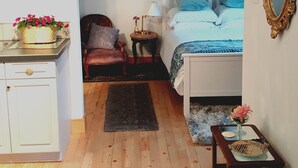 Et soveværelse med trægulv, seng med blåt og hvidt sengetæppe, et natbord med lampe, en stol og en kommode.
