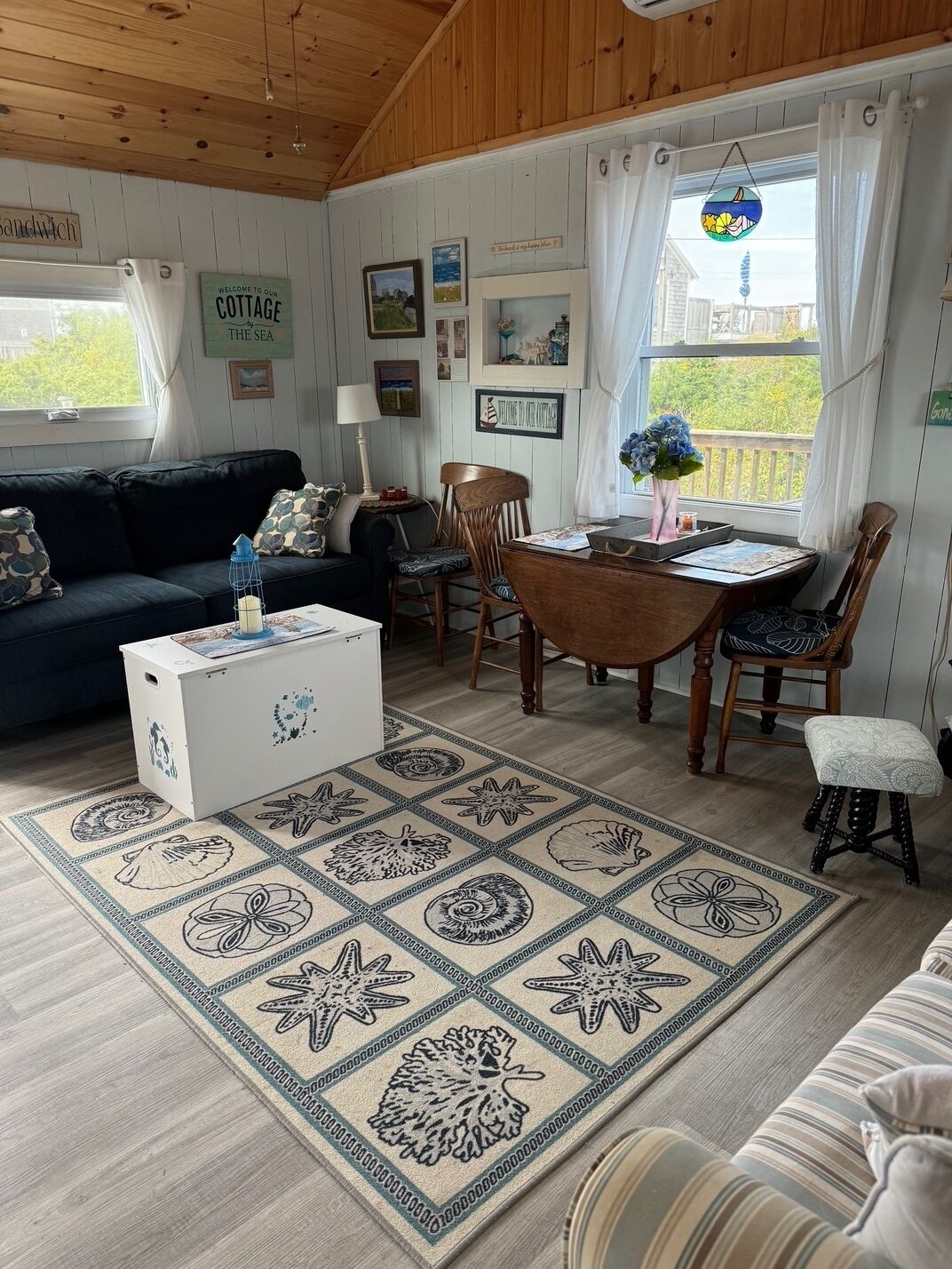 A cozy cabin interior with a wooden ceiling, a dining area with a table and chairs, a sofa, a coffee table, and a rug with sea-themed designs.