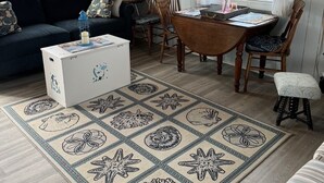 A cozy cabin interior with a wooden ceiling, a dining area with a table and chairs, a sofa, a coffee table, and a rug with sea-themed designs. -  Cozy Cape Cottage on Private Beach with HVAC split AC/Heating system (Sandwich)