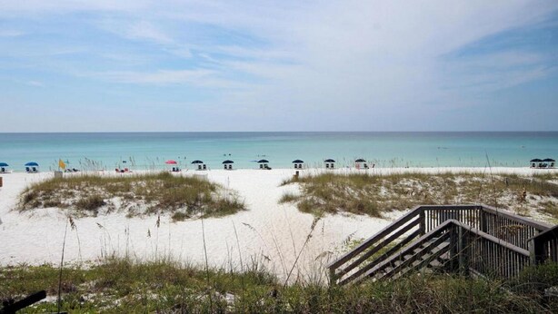 A beach with a wooden staircase leading down to the sand, a row of beach umbrellas, and a clear view of the ocean.