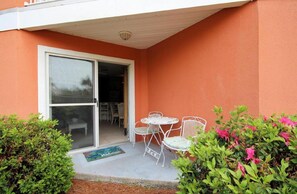 A covered patio with a sliding glass door, white outdoor furniture, and a small garden area with pink flowers. - Beautiful Ground Floor Unit! (Miramar Beach)