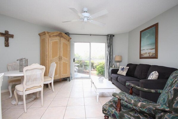 A living room with a white ceiling fan, a dining area with a white table and chairs, a black sofa with patterned cushions, and a large window with a view of greenery. - Beautiful Ground Floor Unit! (Miramar Beach)