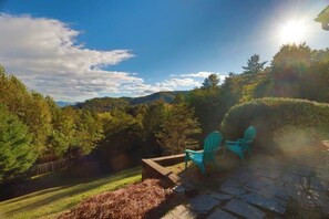 Eine Terrasse im Freien mit Steinböden und zwei grünen Stühlen, umgeben von einem üppigen Garten und Blick auf ferne Hügel.