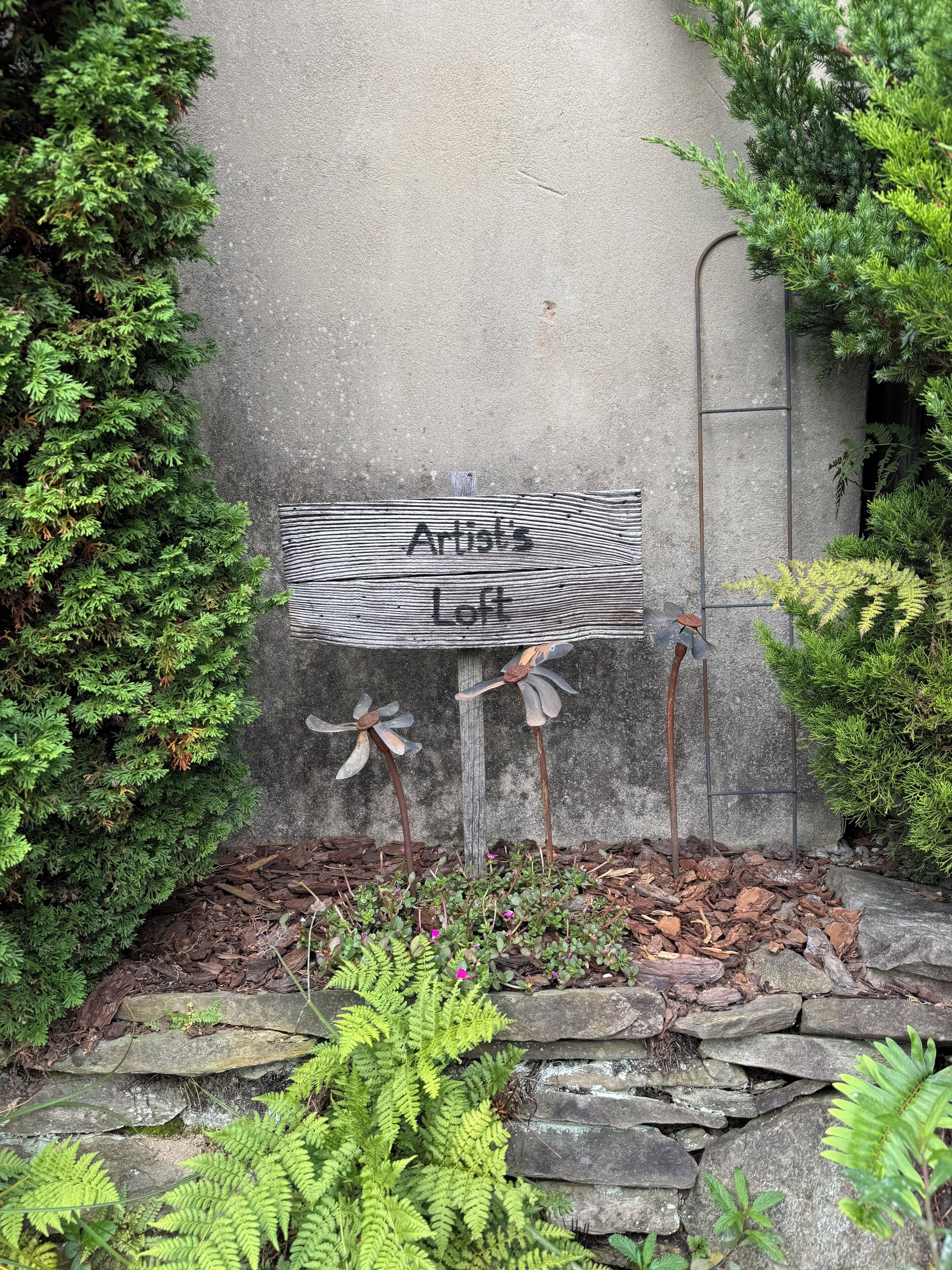 Un letrero de madera rústico con la inscripción "Artist's Loft" sobre un muro de hormigón, rodeado de abundante follaje y un sendero de piedra.