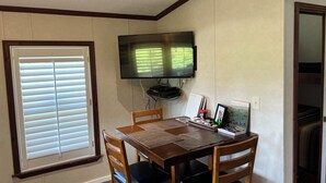 Un coin repas comprenant une table en bois, quatre chaises, un téléviseur fixé au mur et une fenêtre avec des stores.