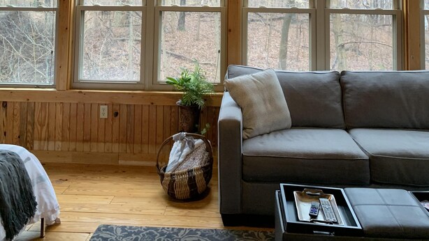 A cozy cabin interior with a ceiling fan, large windows, a gray sofa, and a basket with items.