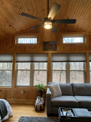 A cozy cabin interior with a ceiling fan, large windows, a gray sofa, and a basket with items.