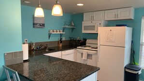 Cozinha com bancadas de granito, armários brancos, micro-ondas, fogão e geladeira. Há duas luminárias pendentes, uma pia e uma lixeira.