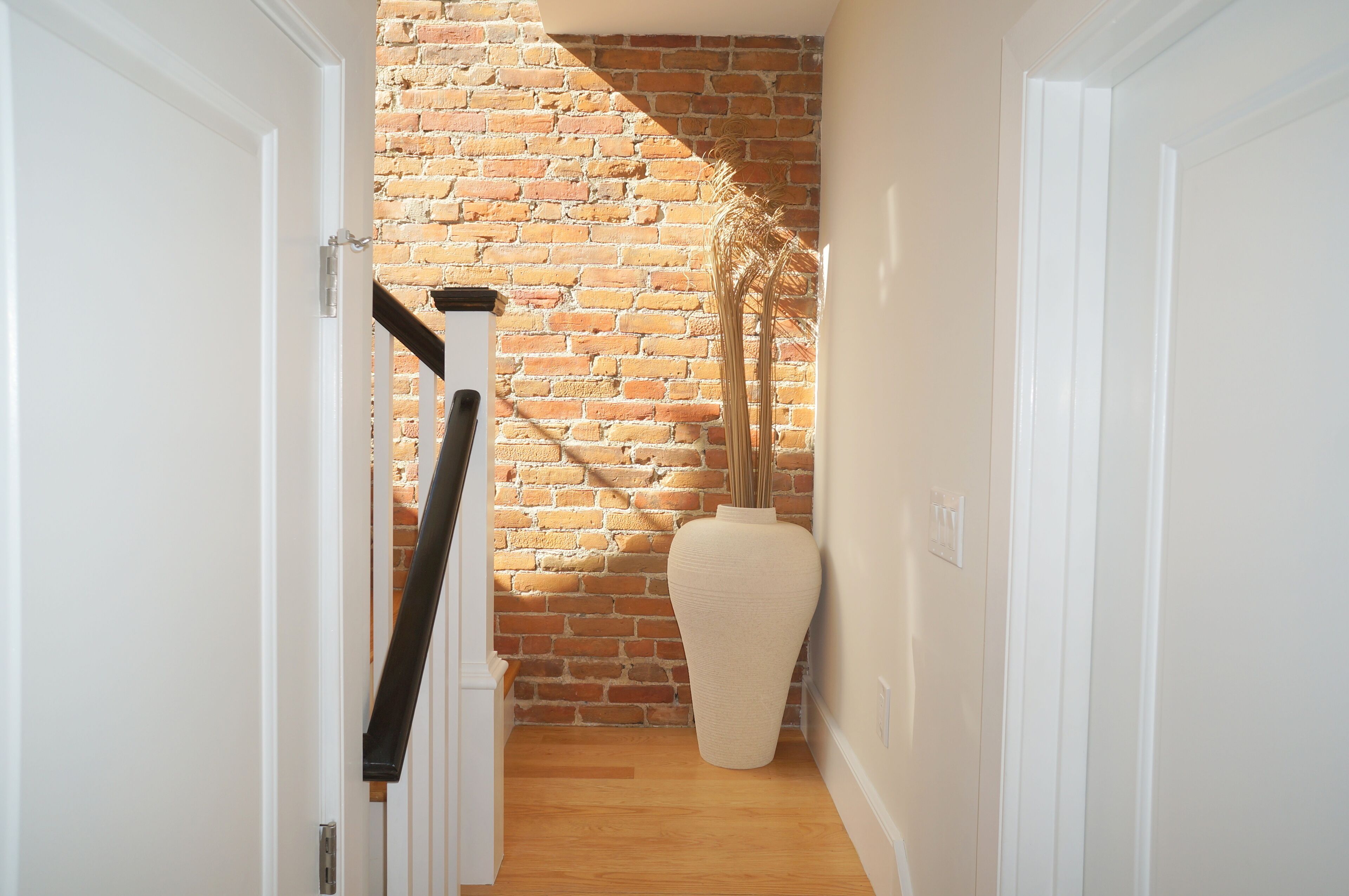 Un pasillo estrecho con una pared de ladrillo, una puerta blanca y un jarrón grande con plantas secas.