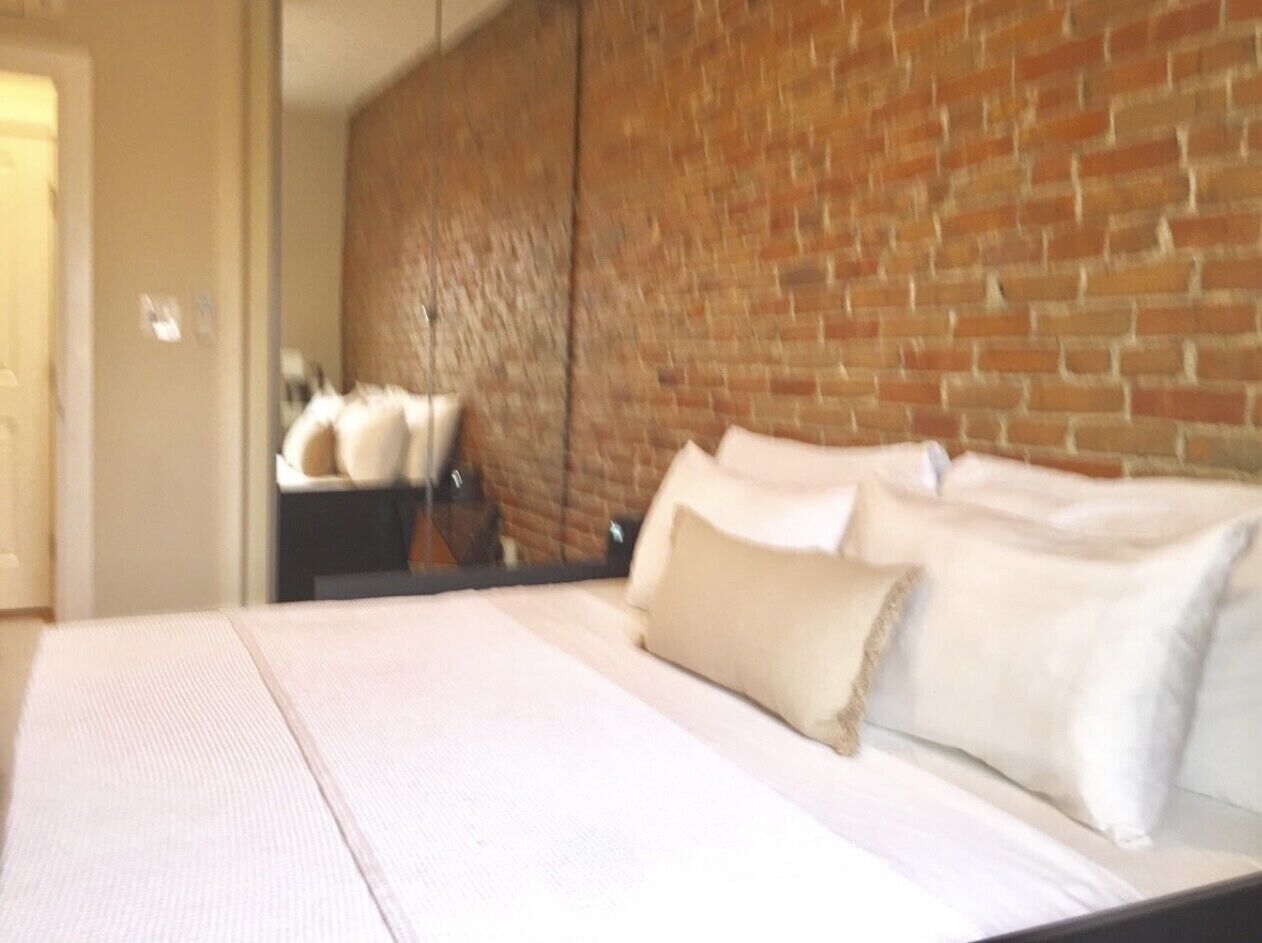 A hotel room with a brick wall, a bed with white linens, and a nightstand with a lamp.