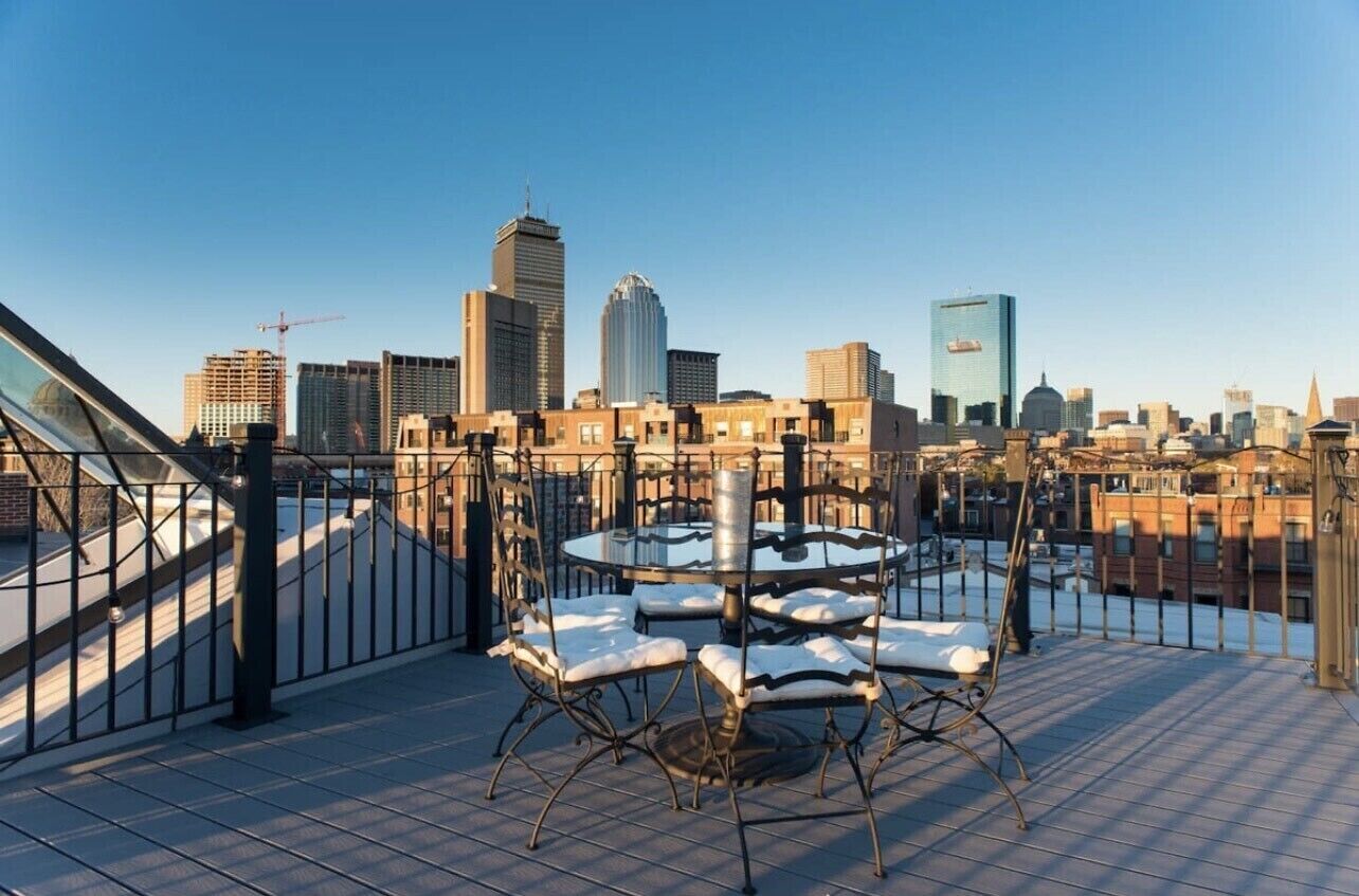 A rooftop terrace with a round table and chairs, overlooking a city skyline with various buildings.
