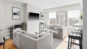 Uma sala de estar moderna com um sofá seccional cinza, uma mesa de centro, uma TV fixada na parede e uma grande janela que oferece luz natural.