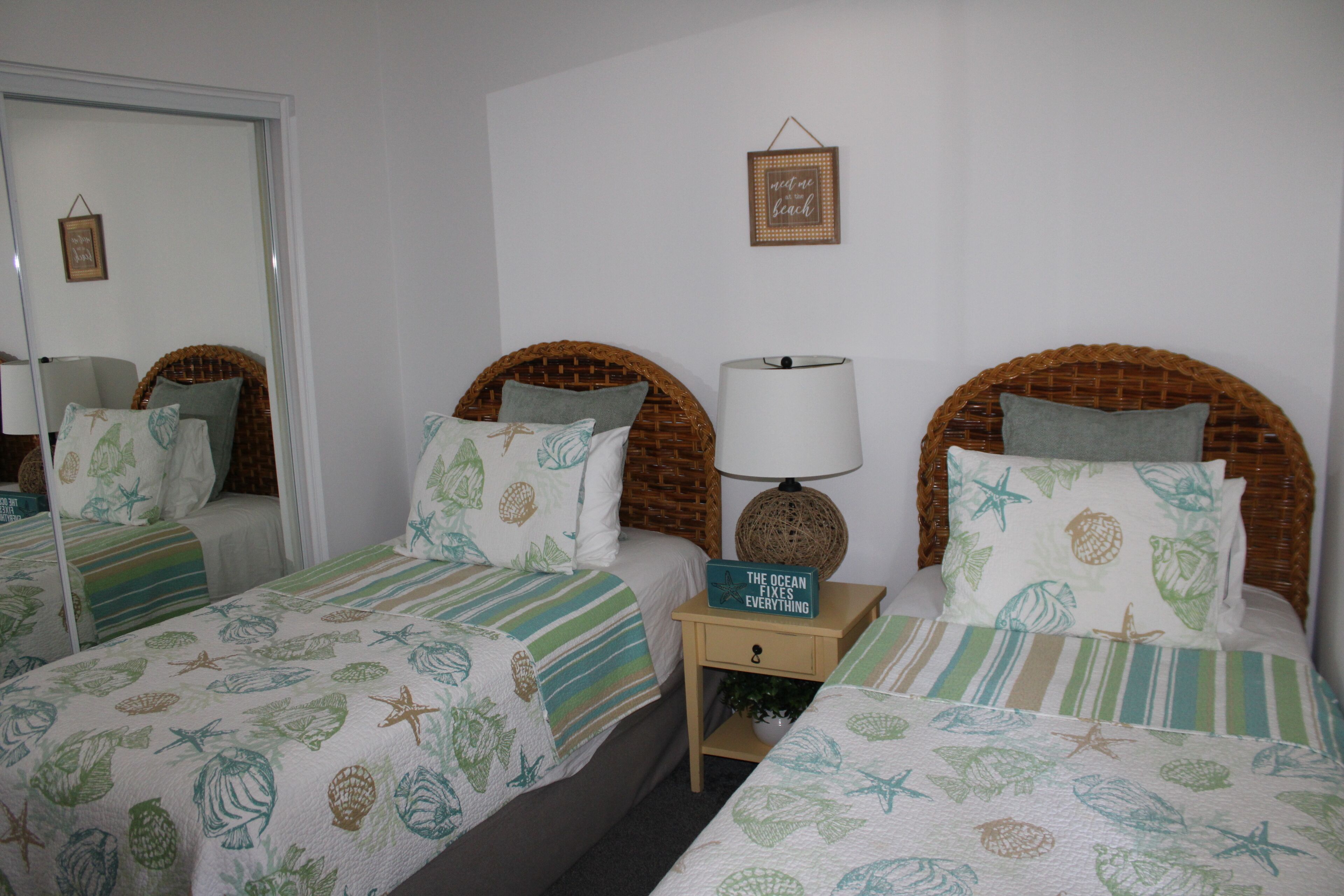 A room with two beds, each with a wicker headboard and a striped bedspread featuring sea creatures. There is a small bedside table with a lamp and a sign that reads "The Ocean Fixes Everything."