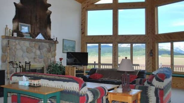 A spacious living room with a stone fireplace, a large wooden sculpture, a television, and a view of a mountain landscape through large windows.