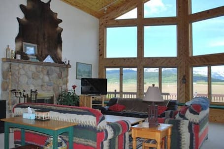 A spacious living room with a stone fireplace, a large wooden sculpture, a television, and a view of a mountain landscape through large windows.