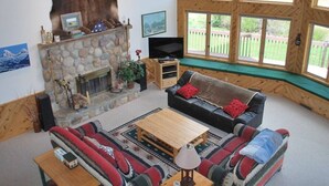 A cozy living room with a stone fireplace, a leather sofa, a wooden coffee table, and large windows offering a view of the outdoors.