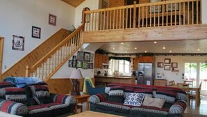 A spacious living area with a high ceiling, wooden staircase, and a kitchen in the background.