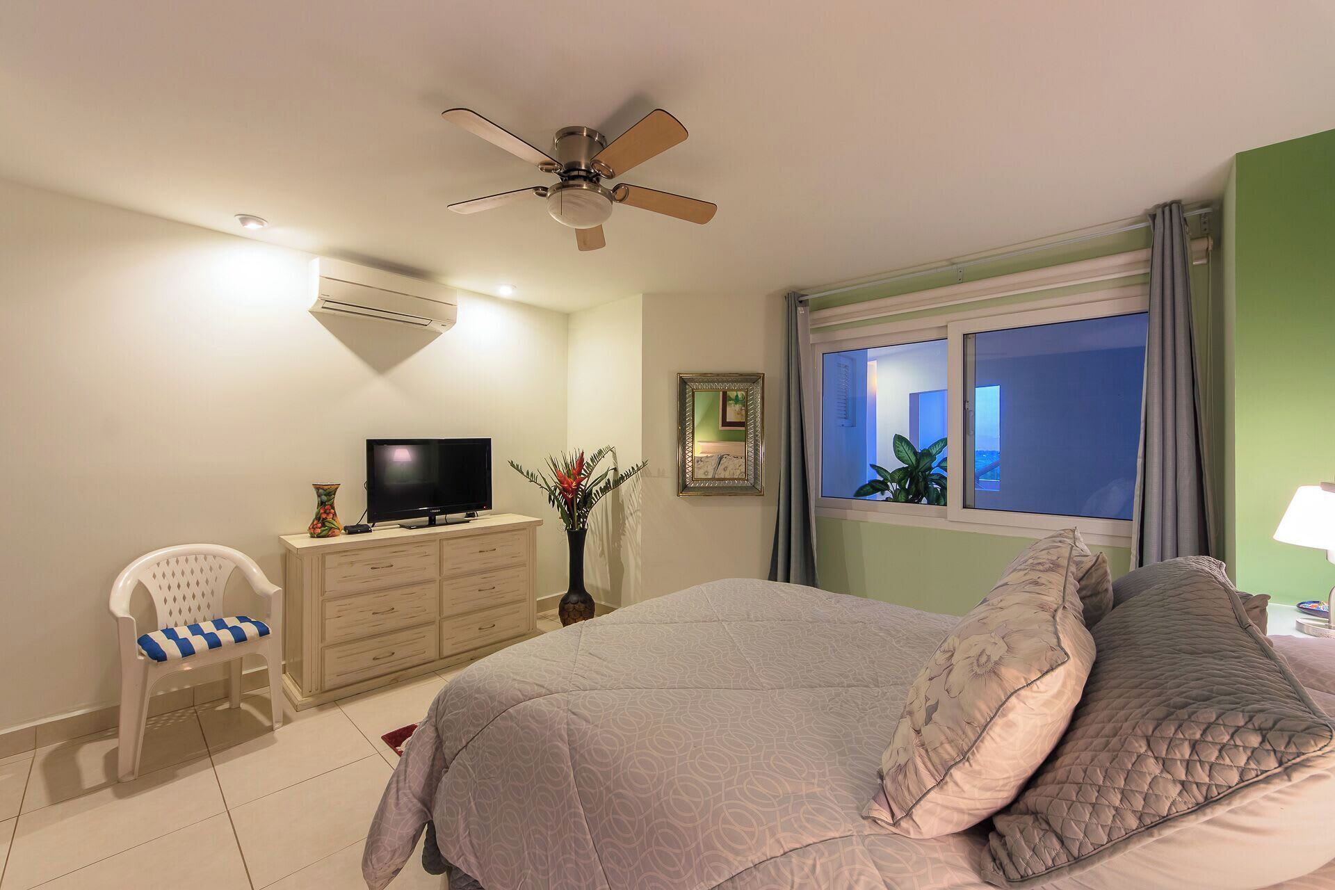 Ein Schlafzimmer mit Bett, Fernseher, Sessel, Kommode, Deckenventilator und einem Fenster mit Aussicht.