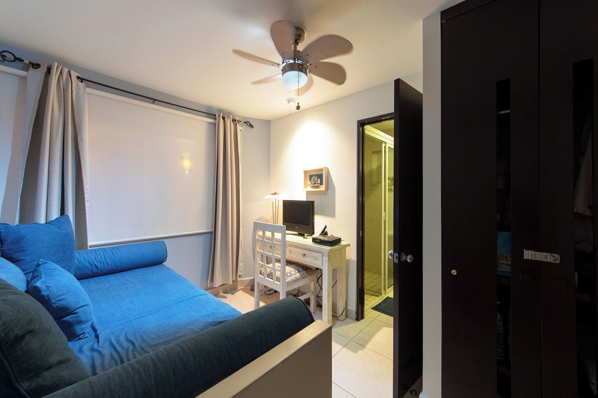 A bedroom with a bed, a desk with a computer, a ceiling fan, and a door leading to another room.