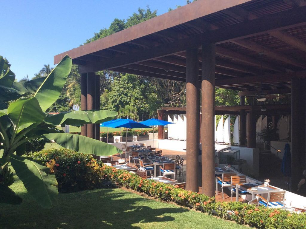 An outdoor patio area with tables and chairs, blue umbrellas, and a covered structure with columns.