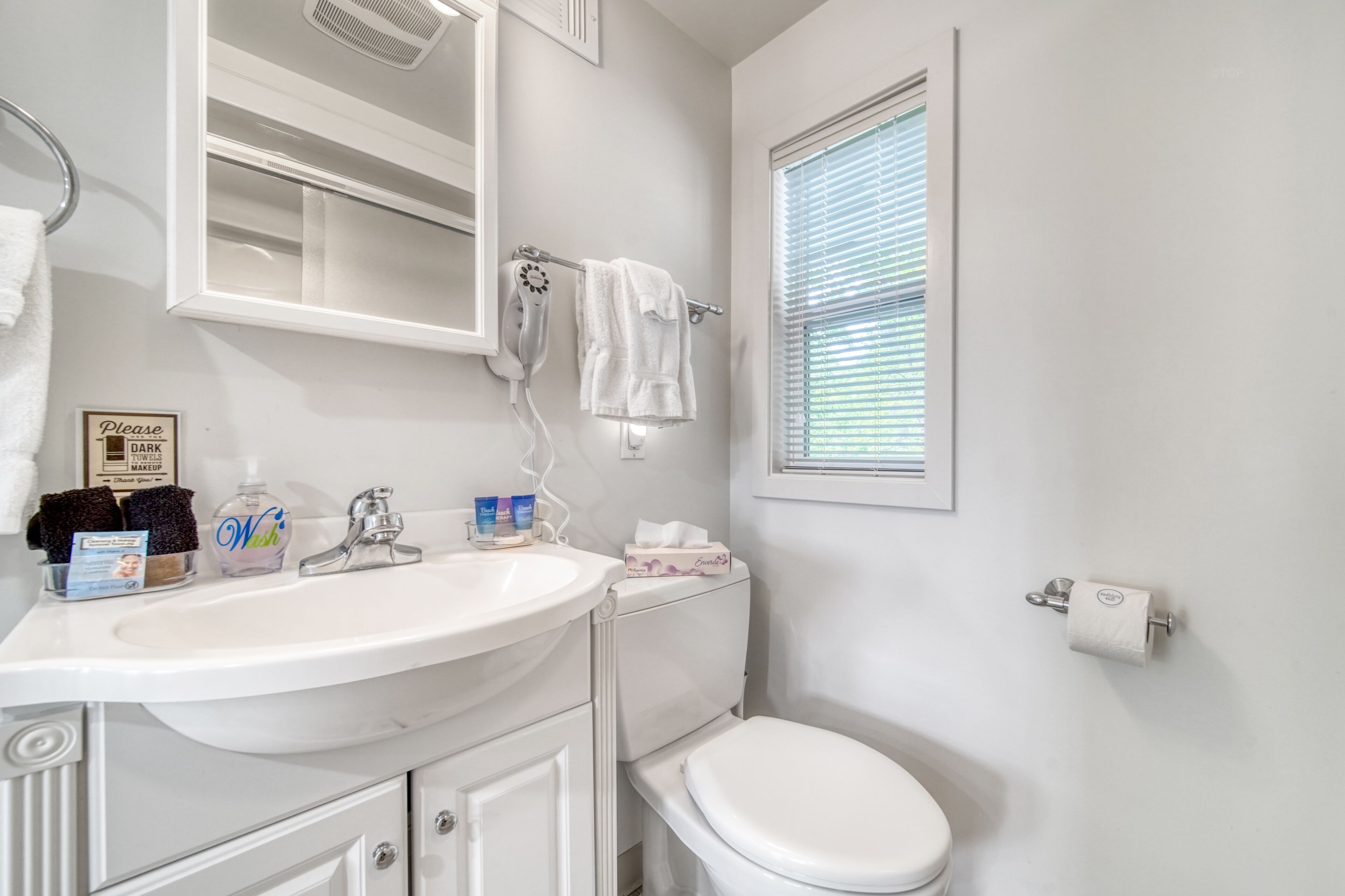 A bathroom with a white sink, mirror, and toilet. There is a towel rack with towels, a bottle of soap, and a box of tissues.