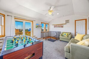 Une pièce avec une table de baby-foot, un canapé, un ventilateur de plafond et une vue sur les maisons à travers de grandes fenêtres.