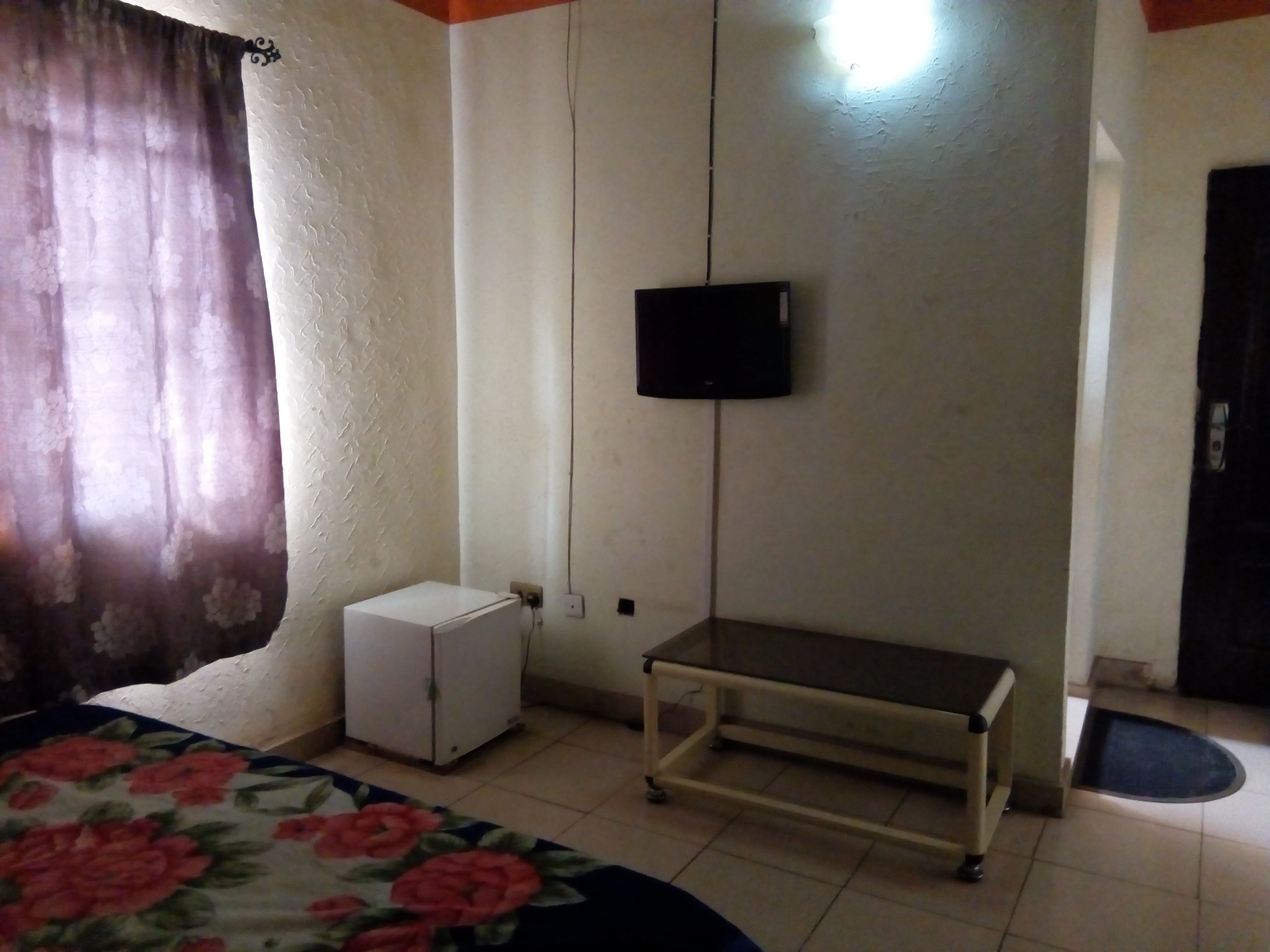 Una stanza modesta con un letto, un frigorifero bianco, una TV a parete e un piccolo tavolo.