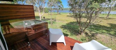Een houten terras met een jacuzzi en witte stoelen, met uitzicht op een gazon met bomen en een waterpartij.