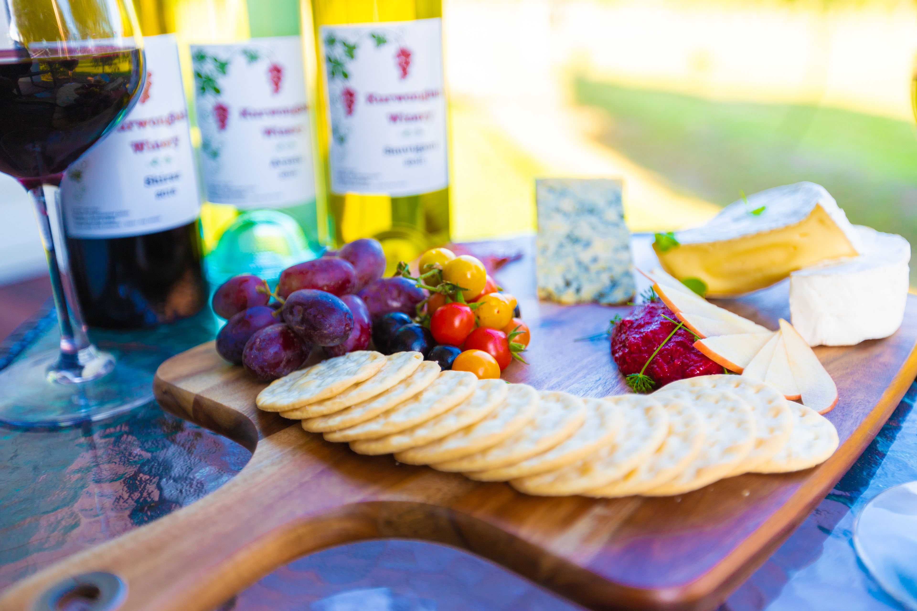A wooden board with cheese, crackers, grapes, and tomatoes, along with wine bottles and a glass of red wine.