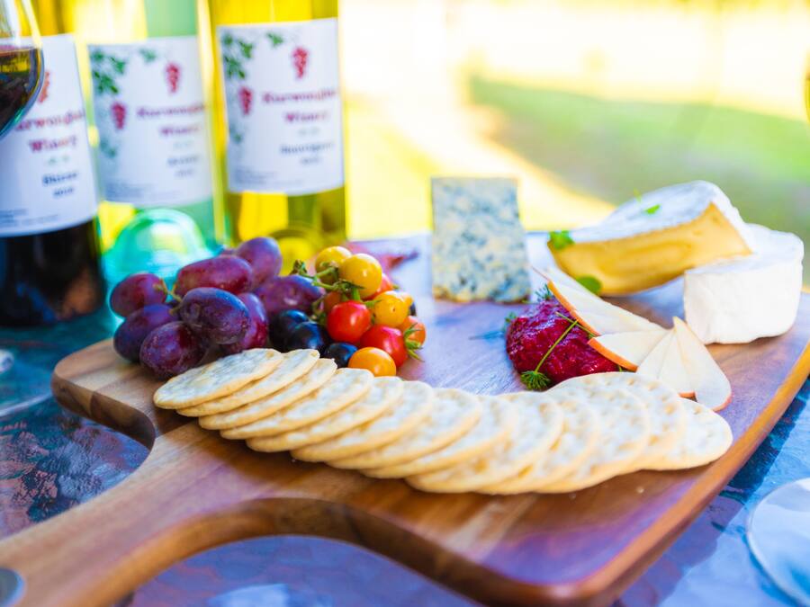 Una tabla de madera con queso, galletas, uvas y tomates, junto con botellas de vino y una copa de vino tinto.