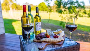 Une table en osier, placée à l’extérieur, sur laquelle on trouve des bouteilles de vin, un verre de vin rouge, du fromage, des raisins et des crackers.