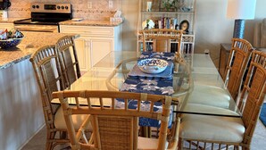 A dining area with a glass table and wicker chairs, a kitchen with modern appliances, and a decorative shelf with books and plants. - Dir Ocean Front 2 BR/2 BA Condo (ST AUGUSTINE)