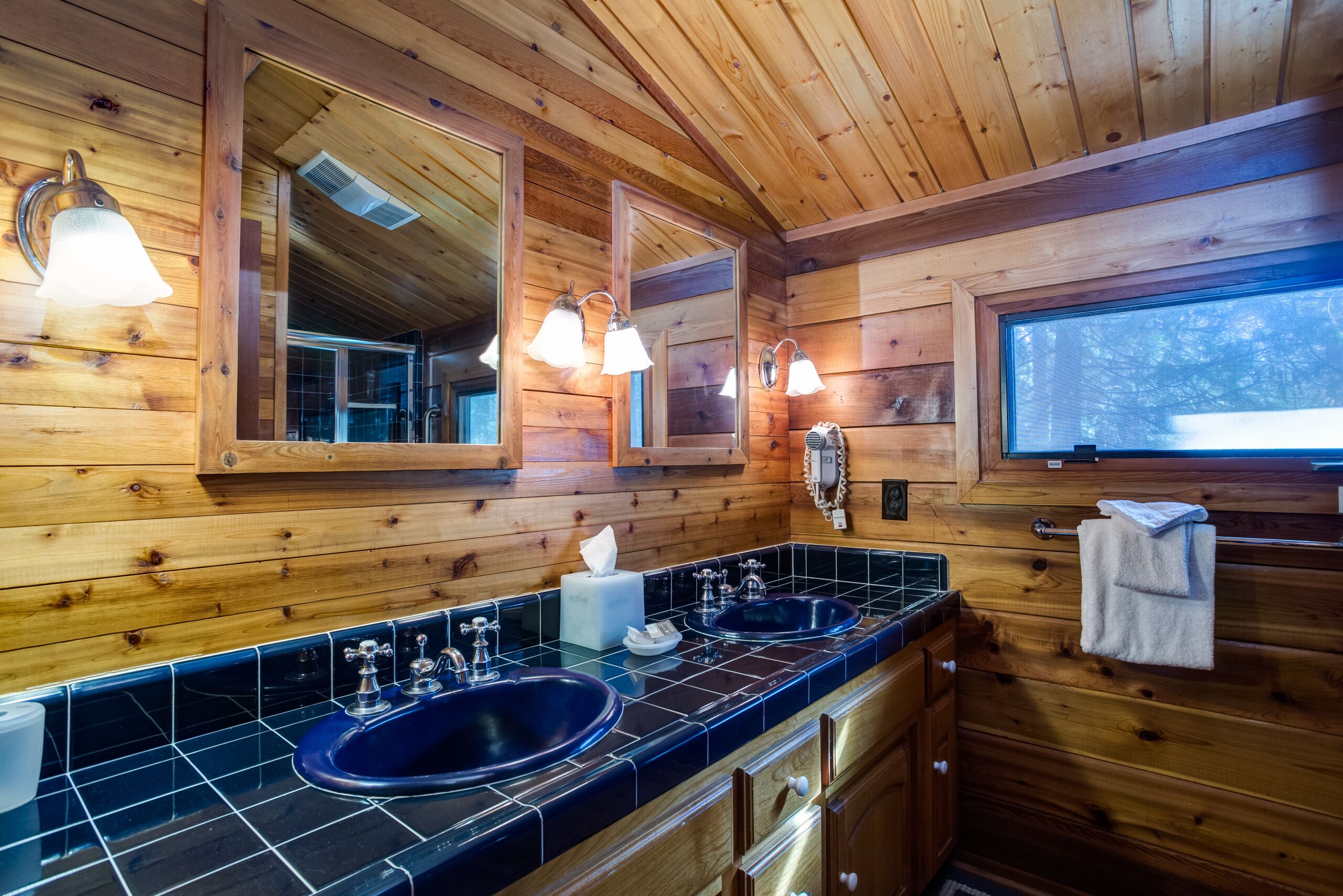Een houten badkamer in een vakantiehuis, met twee wastafels, een spiegel en een raam.