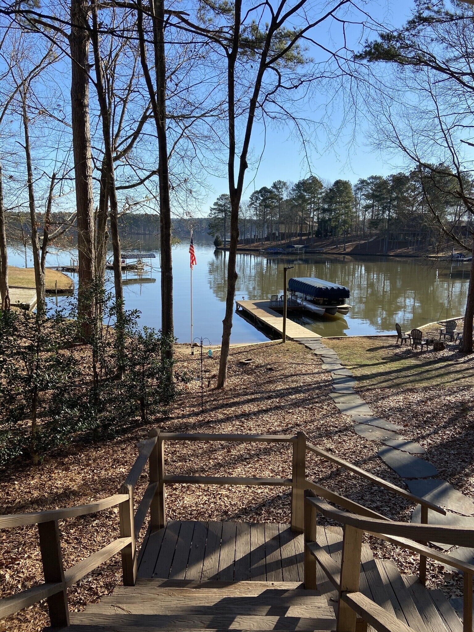 A wooden deck with a view of a calm lake, surrounded by trees and a dock with a small boat.