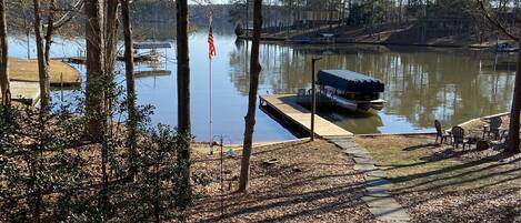 A wooden deck with a view of a calm lake, surrounded by trees and a dock with a small boat.