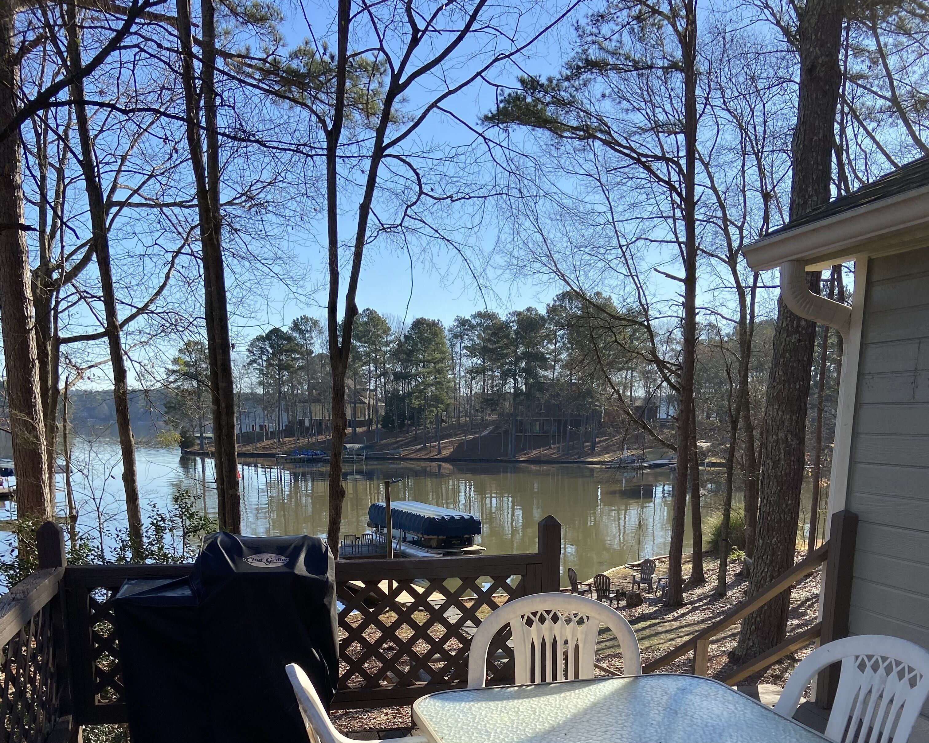 A lakeside deck with a grill, chairs, and a view of the lake and surrounding trees.