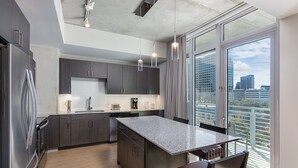 A modern kitchen with dark cabinets, a granite countertop, and a city view through large windows. - Austin | Couples ATX Base | Pool + Gym (Austin)
