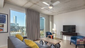 A modern living room with a grey sofa, yellow pillows, a coffee table with wheels, a TV, and a city view through large windows. - Austin | Couples ATX Base | Pool + Gym (Austin)