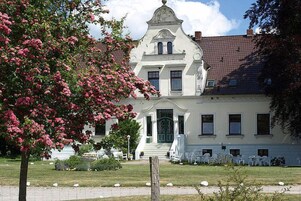 Um edifício branco com porta verde, rodeado por um jardim com flores cor-de-rosa e cadeiras brancas.