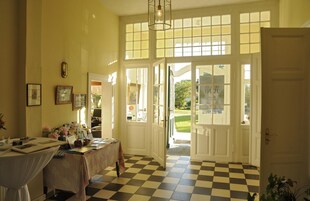 Um quarto com chão quadriculado, uma mesa com diversos objetos e uma porta que dá para o exterior.