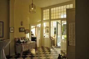 Um quarto com chão xadrez, uma mesa com diversos objetos e uma porta que dá para o exterior.